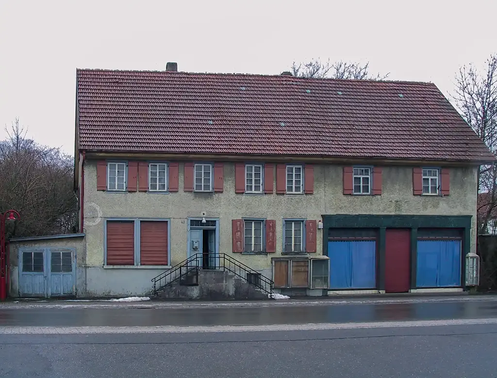 Ein altes Gebäude mit großen Schaufenstern und roten Fensterläden. Vor dem alten Kaufhausgebäude ist eine kleine Straße.