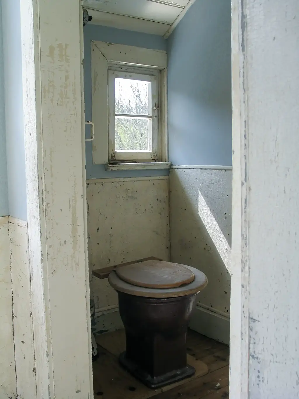 Ein einfaches, altmodisches WC mit blauen Wänden. Ein kleines Fenster lässt natürliches Licht hinein.