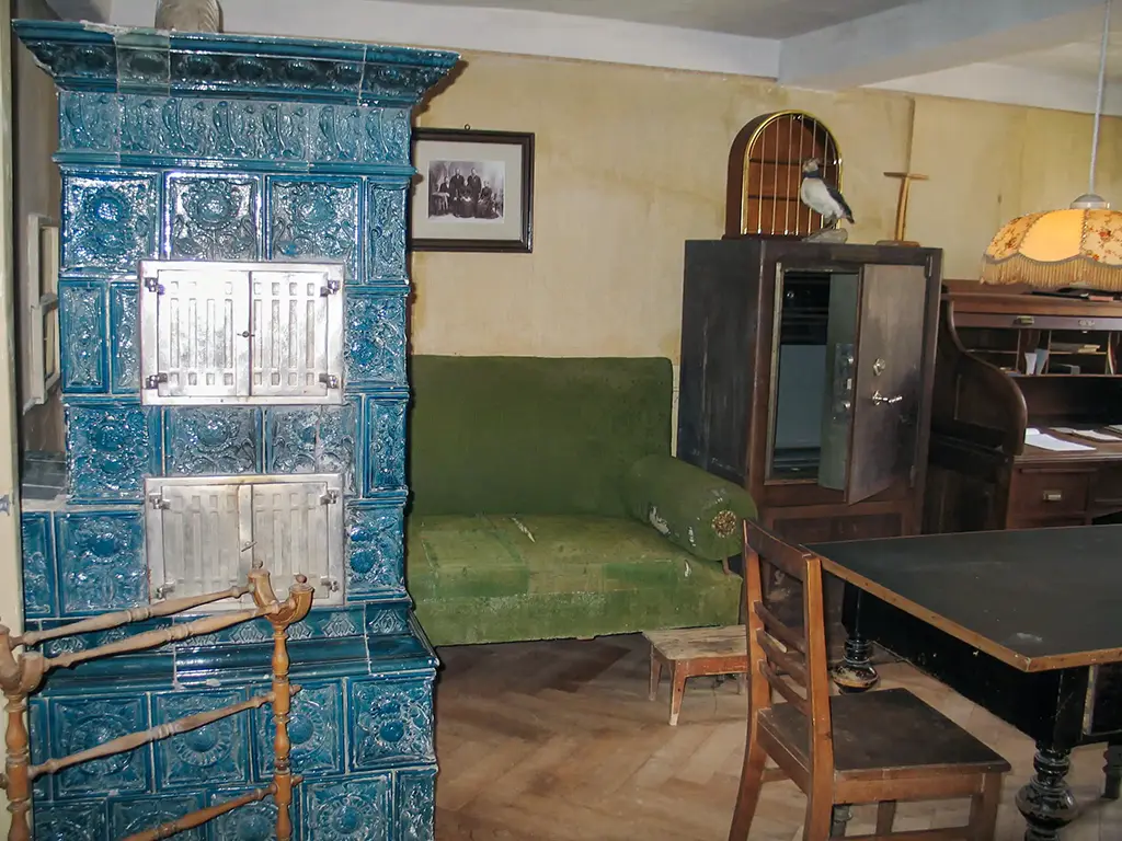 Wohnraum mit einem großen, verzierten Kachelofen in Blau, einer grünen Couch und einem Tisch mit Stühlen.