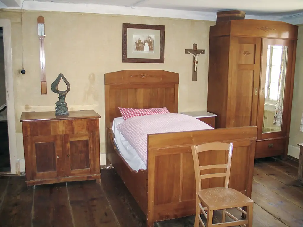 Schlafzimmer mit Einzelbett, drauf mit rot-weiß karierte Bettwäsche. Außerdem sind in dem Zimmer ein Schrank, ein kleiner Nachttisch und einem Wandkreuz, das eine einfache, rustikale Atmosphäre schafft.