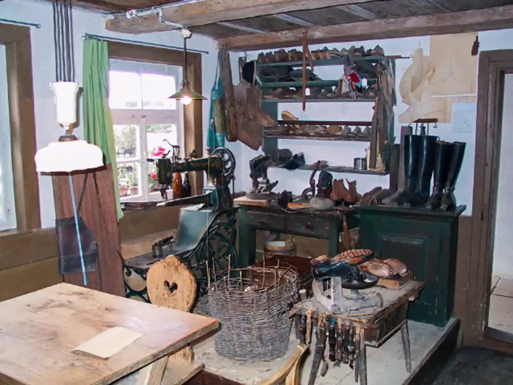 Eine mit Holzmöbeln eingerichtete Schusterwerkstatt mit Nähmaschine und Werkzeugen. In einem Regal an der Wand und im Raum verteilt liegen viele Schuhe.