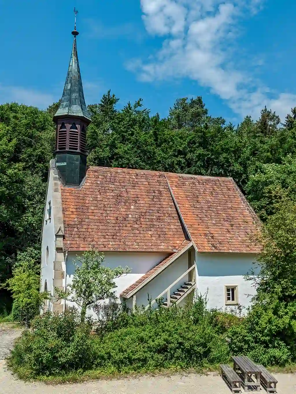 Seitliche Außenansicht der Dorfkirche Tischardt mit Treppe zur Empore, rote Ziegeldach und bewaldeter Hintergrund.