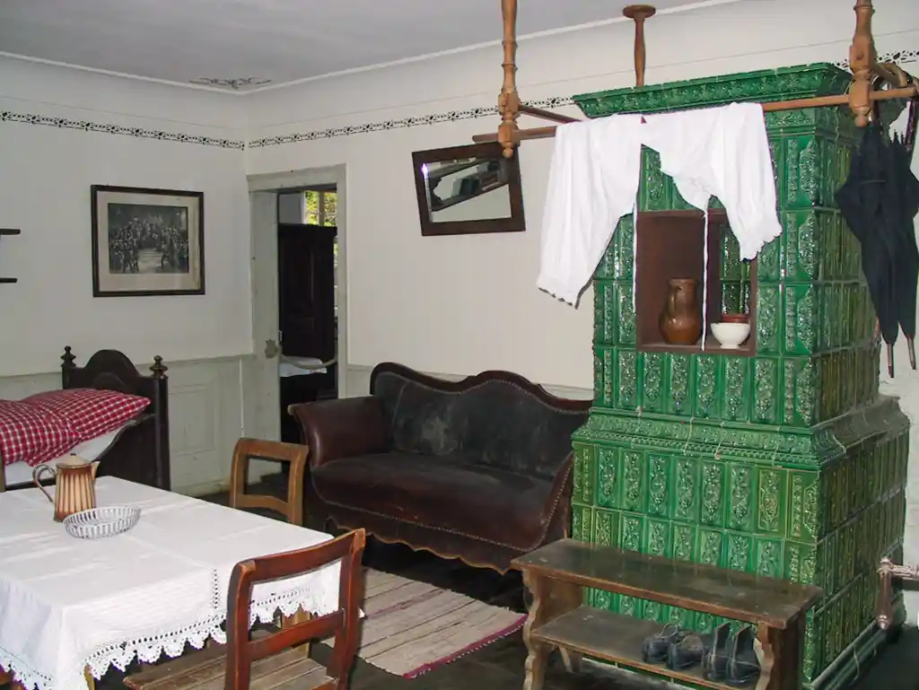 Wohnzimmer mit grünem Kachelofen, Sofa und Esstisch mit Tischdecke.