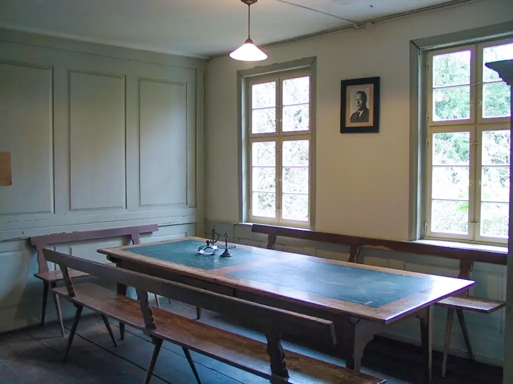 Ein einfacher, langer Tisch mit langen Holzbänken in einem Raum mit weißen Wänden und großen Fenstern. An der Wand hängt ein Porträt.