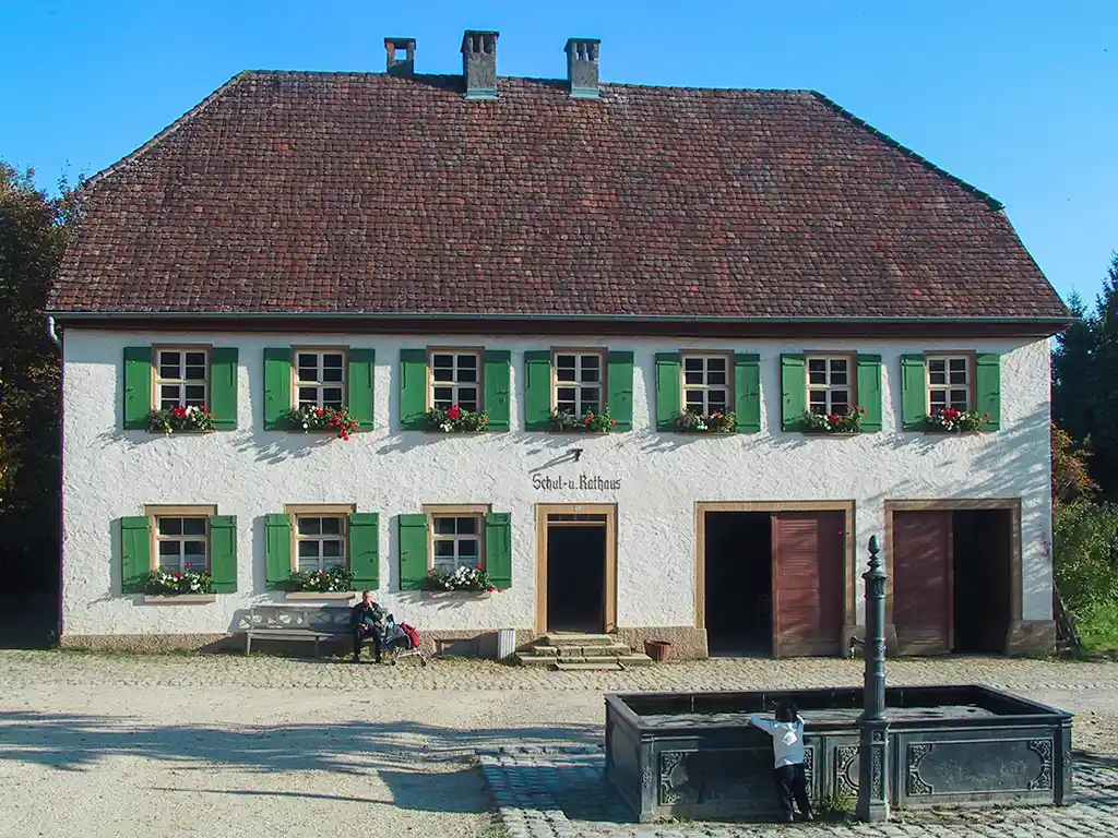 Das historische Schul- und Rathausgebäude mit grün umrahmten Fenstern und Blumenkästen, im Vordergrund ein Brunnen und ein Kind, das am Brunnen spielt.