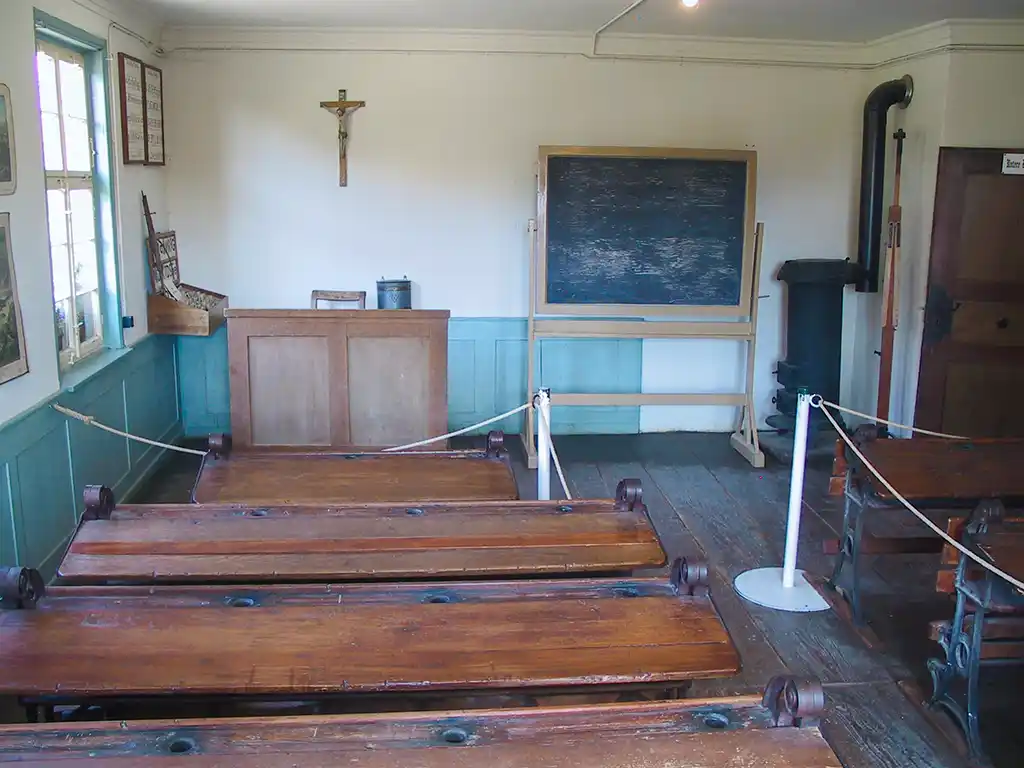 Klassenzimmer mit Holzschulbänken, Tafel und Ofen. Hinter dem Lehrerpult hängt ein religiöses Kreuz an der Wand.