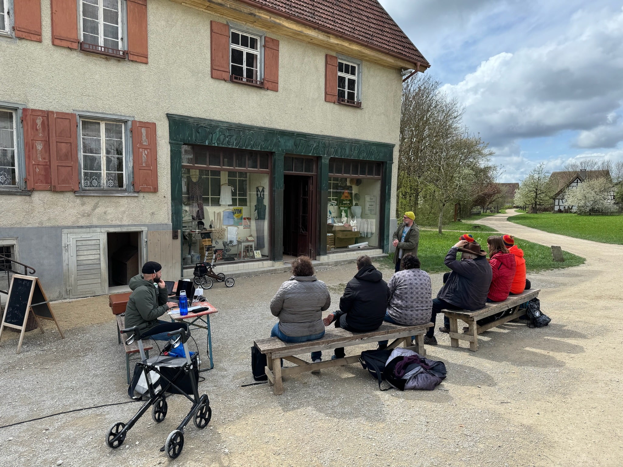 Das Bild zeigt eine kleine Gruppe von Menschen, die vor einem historischen Kaufhaus im Freilichtmuseum auf einfachen Holzbänken sitzt. Eine Person steht vor dem Laden und spricht zur Gruppe. Neben dem Eingangsbereich sitzt eine weitere Person mit Laptop an einem Tisch. Vorne im Bild steht ein Rollator. Die Szene spielt an einem teils sonnigen, teils bewölkten Tag.