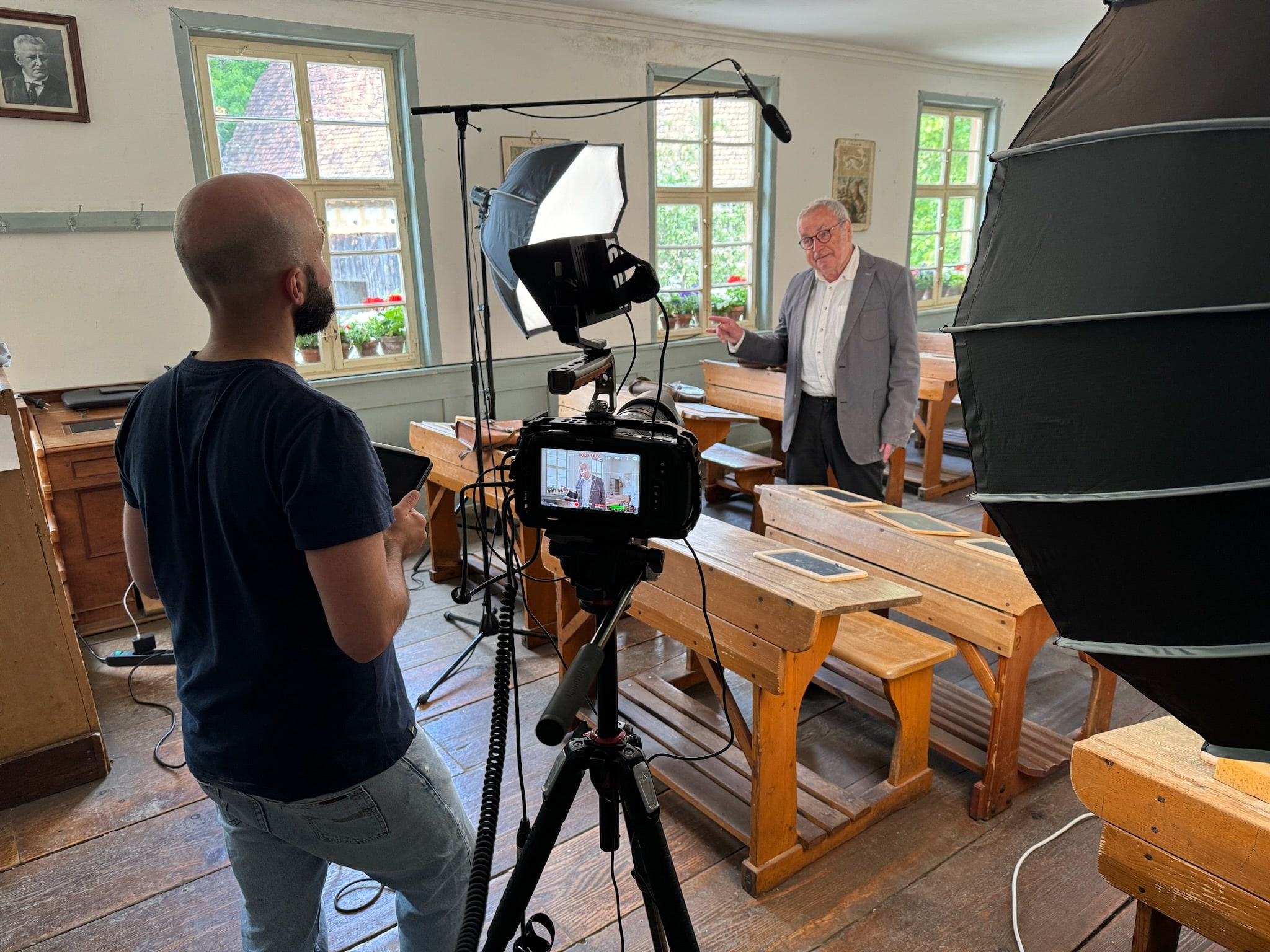 Das Bild zeigt eine professionelle Videoproduktion in einem historischen Klassenzimmer. Ein älterer Mann steht vor alten Holzschultischen und spricht in die Kamera. Eine zweite Person mit Tablet betreut die Aufnahme. Im Vordergrund sind Kamera, Licht- und Tontechnik sichtbar, die auf Stativen aufgebaut sind. Durch die Fenster fällt Tageslicht auf die Szene.
