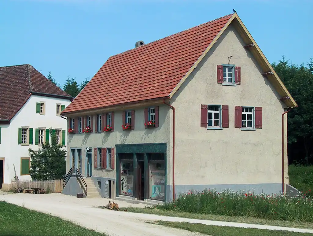 Historisches Gebäude mit Blumen an den Fenstern und roten Fensterläden. Angrenzend ist ein großes Gebäude im Hintergrund.