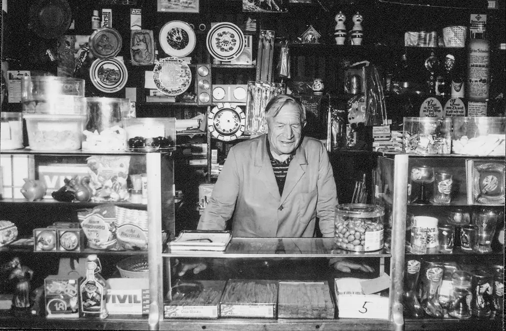 Schwarz-weiß Foto von Franz Pfeiffer, ein älterer Mann, hinter der Ladentheke seines Kaufhauses, umgeben von Regalen mit verschiedenen Produkten.