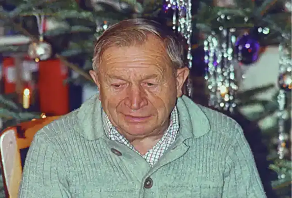 Porträt eines älteren Mannes in einem grauen Pullover vor einem festlich geschmückten Weihnachtsbaum.