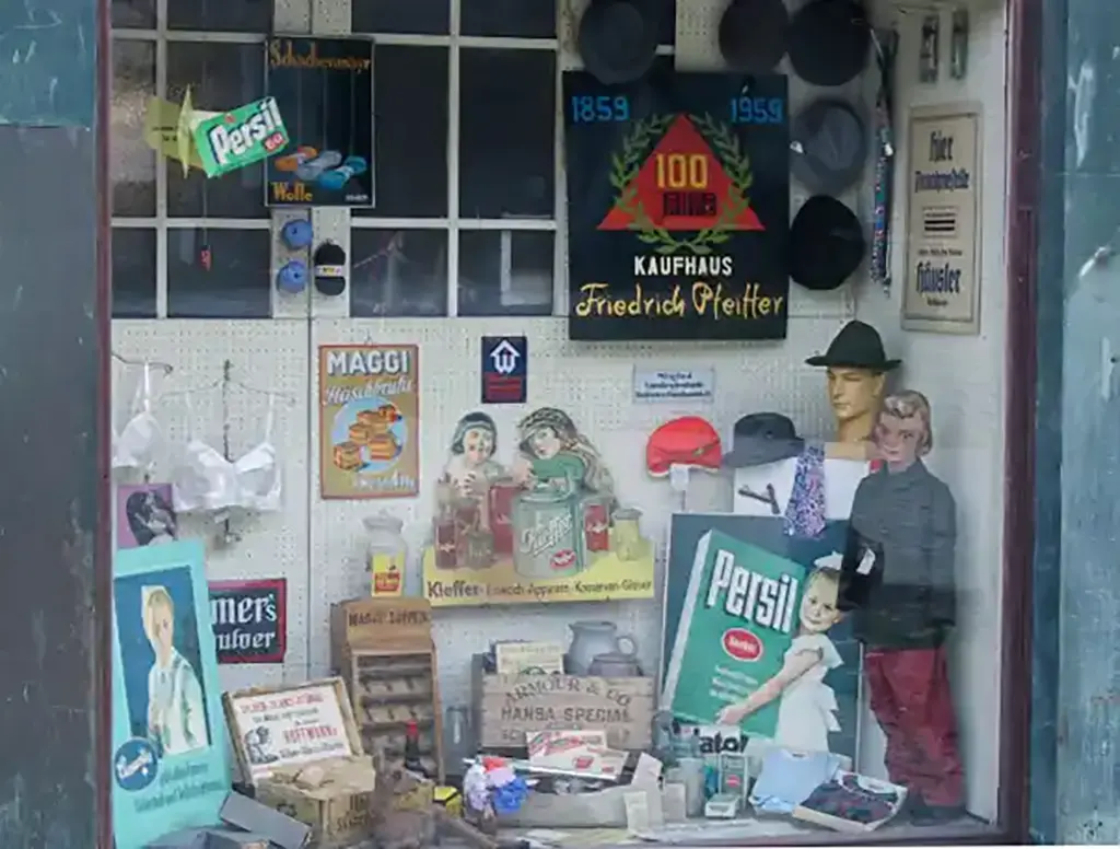 Ein Schaufenster mit nostalgischen Werbeplakaten und Haushaltsartikeln, darunter ein Schild mit dem Namen