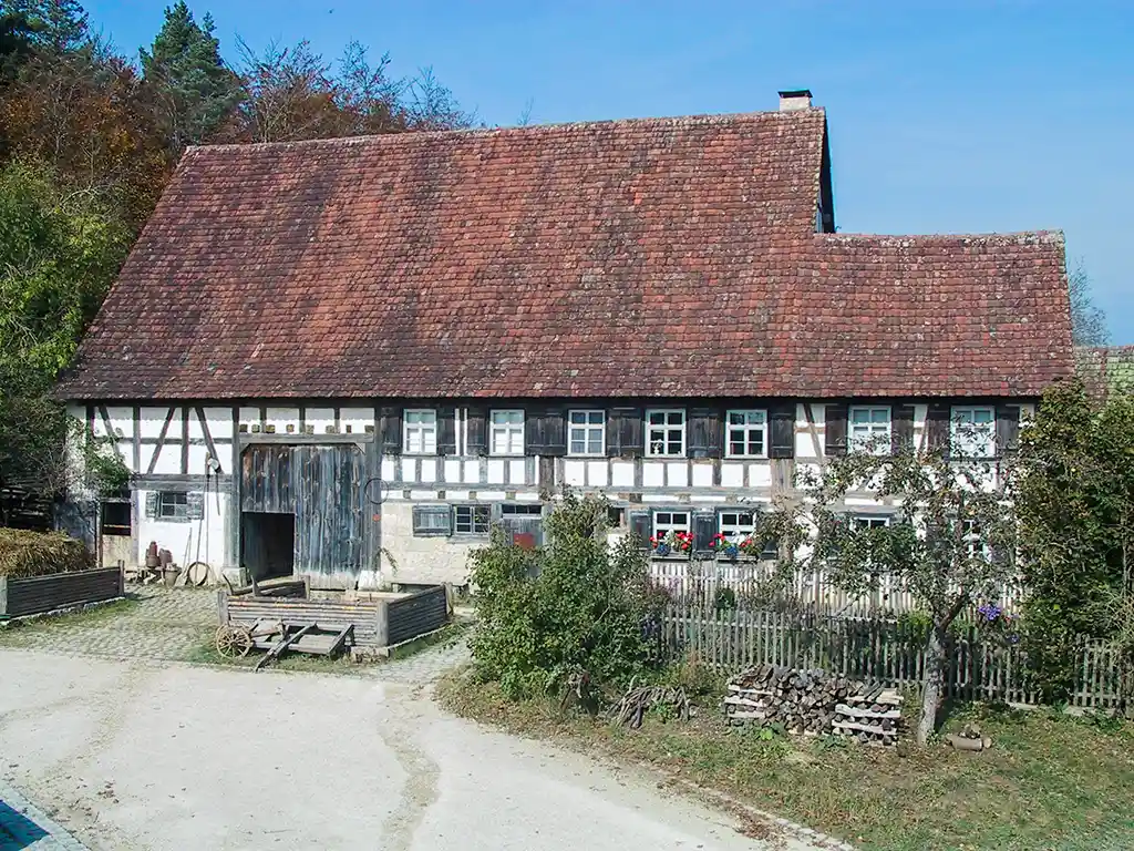 Ansicht eines großen Fachwerkhauses mit roten Dachziegeln. Um das Haus herum befinden sich Bäume und Büsche.