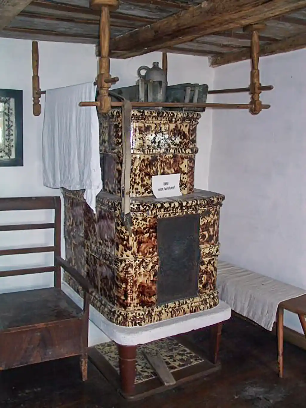 Ein verzierter Kachelofen mit Holzgestell in einem Wohnraum. Über dem Ofen hängen Trockentücher.