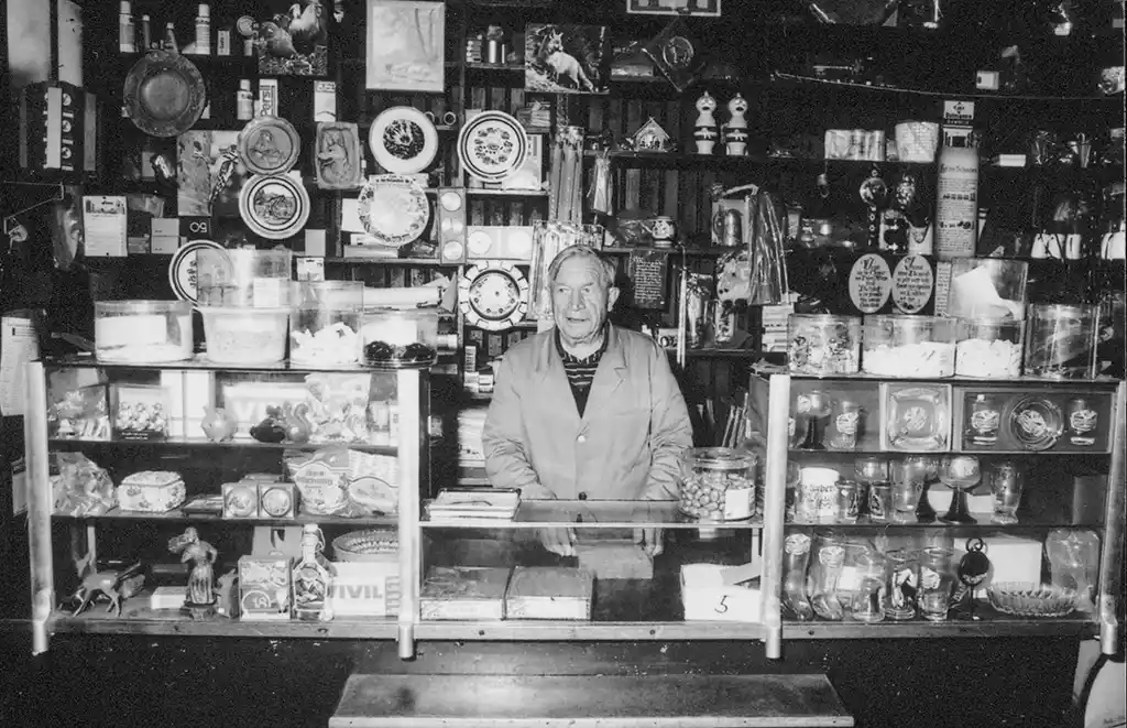 Schwarz-weiß Foto von Franz Pfeiffer, ein älterer Mann, hinter der Ladentheke seines Kaufhauses, umgeben von Regalen mit verschiedenen Produkten.