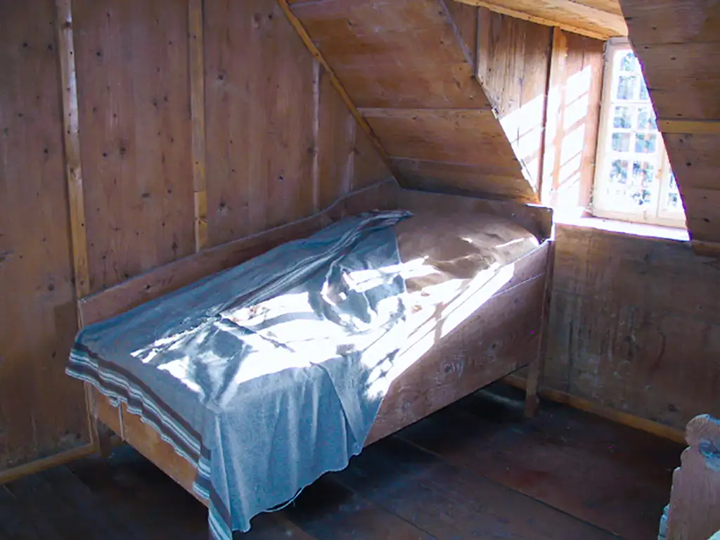 Einfaches Zimmer mit Holzbett unter einer Schräge, kleine Fenster lassen Licht herein.