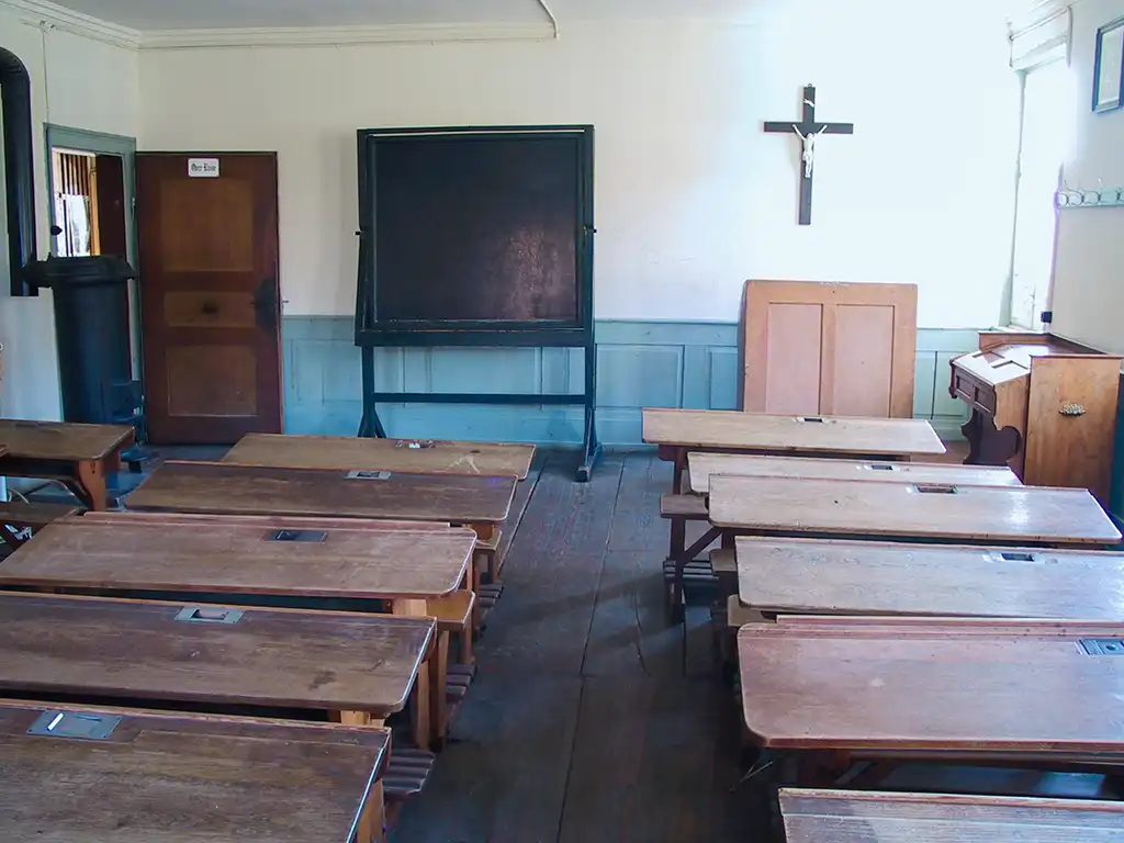 Klassenzimmer mit Holzbänken, großer Schiefertafel und Kreuz an der Wand.