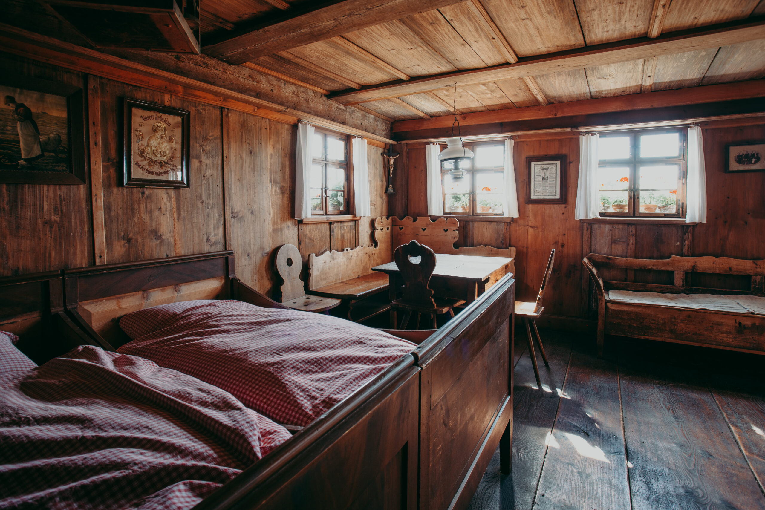 Foto von einem Wohnraum mit Holzwänden und Holzdecke. Im Raum stehen zwei Betten mit karriertem Bettzeug, eine Eckbank, einem Holztisch und Stühlen. Daneben steht eine Holzbank. An den Wänden hängen Bilder und ein Kreuz.