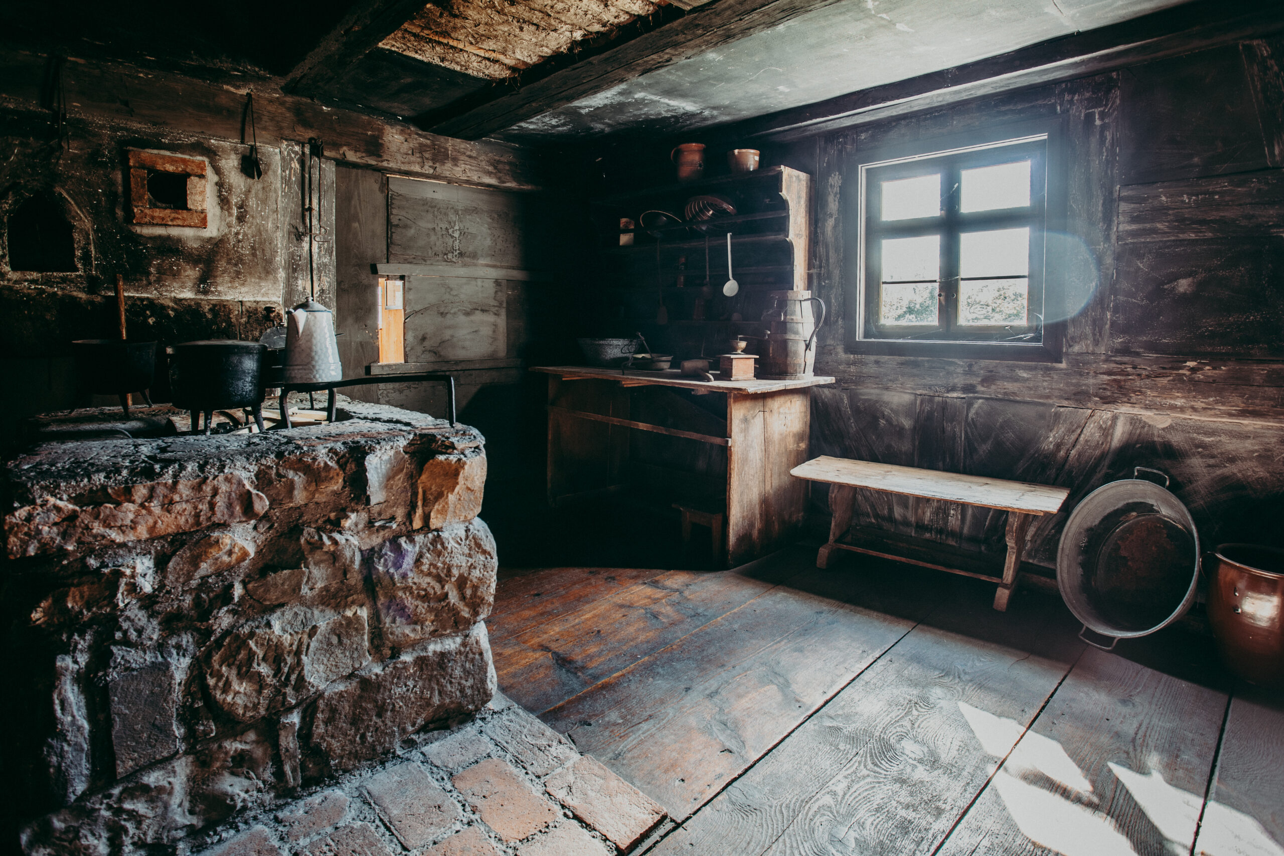 Foto: Innenraum einer historischen Küche mit dunklen Holzwänden und -decke. Links ein gemauerter Herd mit alten Töpfen, rechts ein Holzregal mit Tongefäßen und Küchenutensilien. Sonnenlicht fällt durch ein kleines Fenster