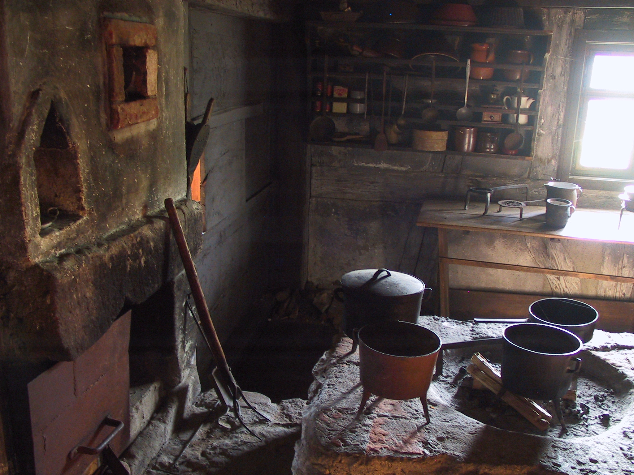 Foto: Historische Küche mit gemauertem Herd, auf dem mehrere schwarze Töpfe stehen. Rechts ein Holztisch mit weiteren Küchenutensilien, darüber ein Regal mit Geschirr, Löffeln und Tongefäßen. Licht fällt durch ein kleines Fenster auf die Szenerie.
