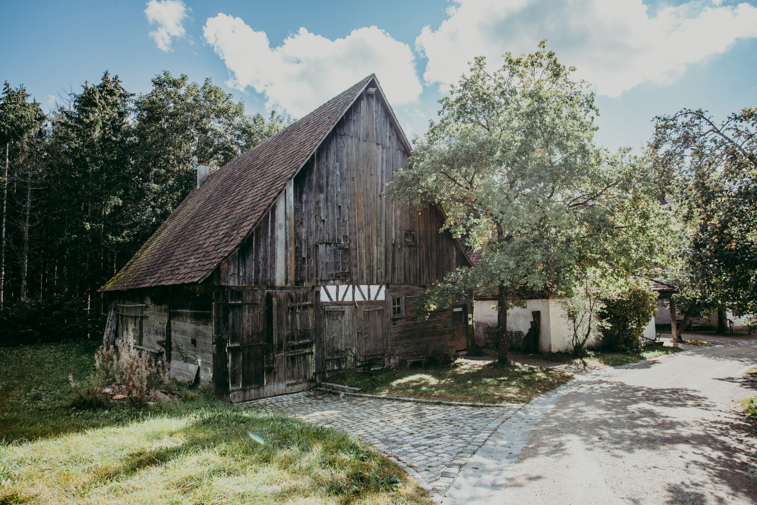 Foto: Außenansicht des Farrenstalls mit hohem, steilem Dach und verwitterter Holzfassade. Der Stall liegt an einem Kopfsteinpflasterweg, umgeben von Bäumen und kleineren Nebengebäuden.