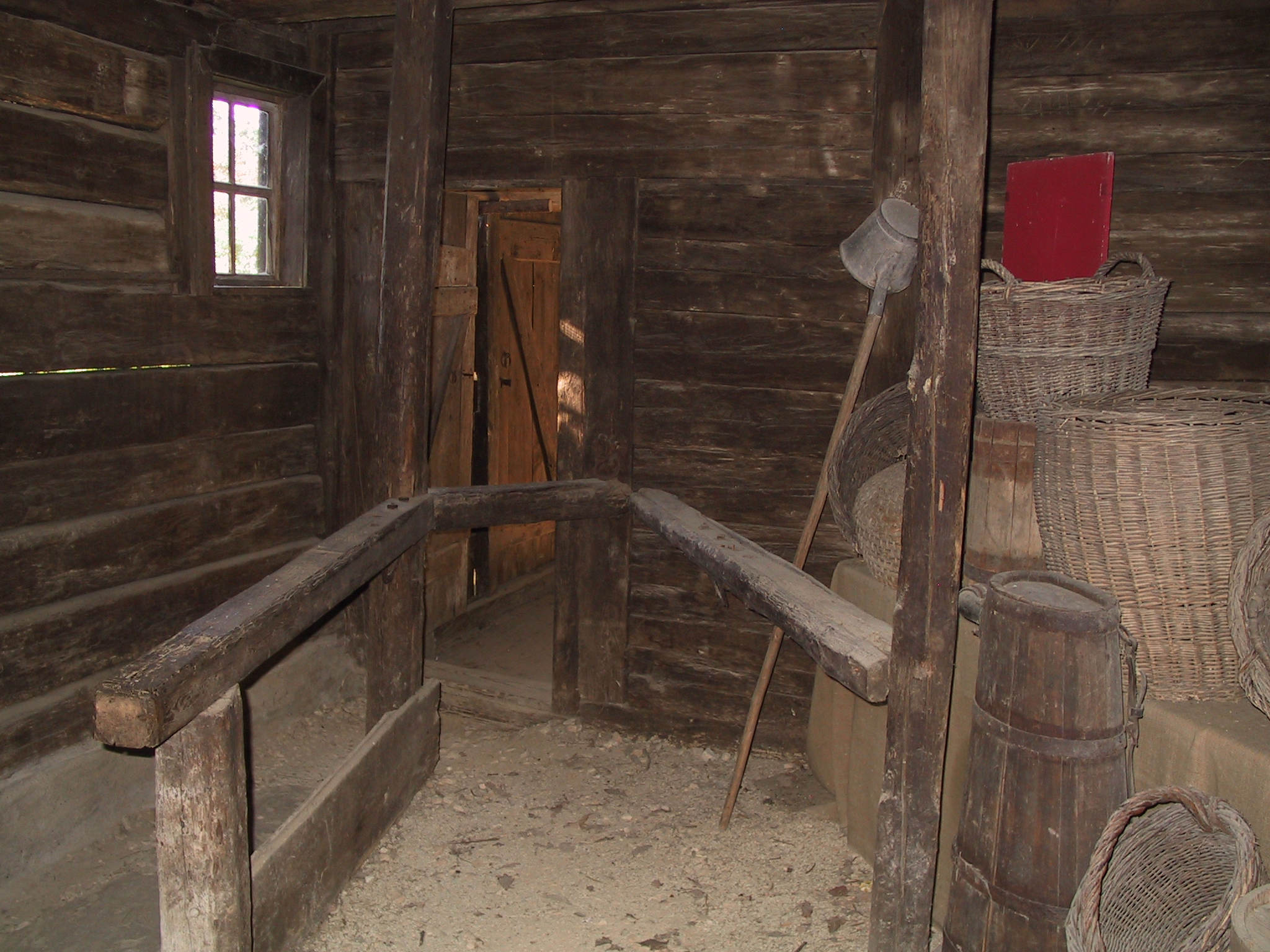 Foto: Abgetrennter Bereich im Inneren eines Farrenstalls. Rechts stehen Körbe auf einem Podest. Licht fällt durch ein kleines Fenster an der linken Wand, die Tür zum anliegenden Raum steht offen.