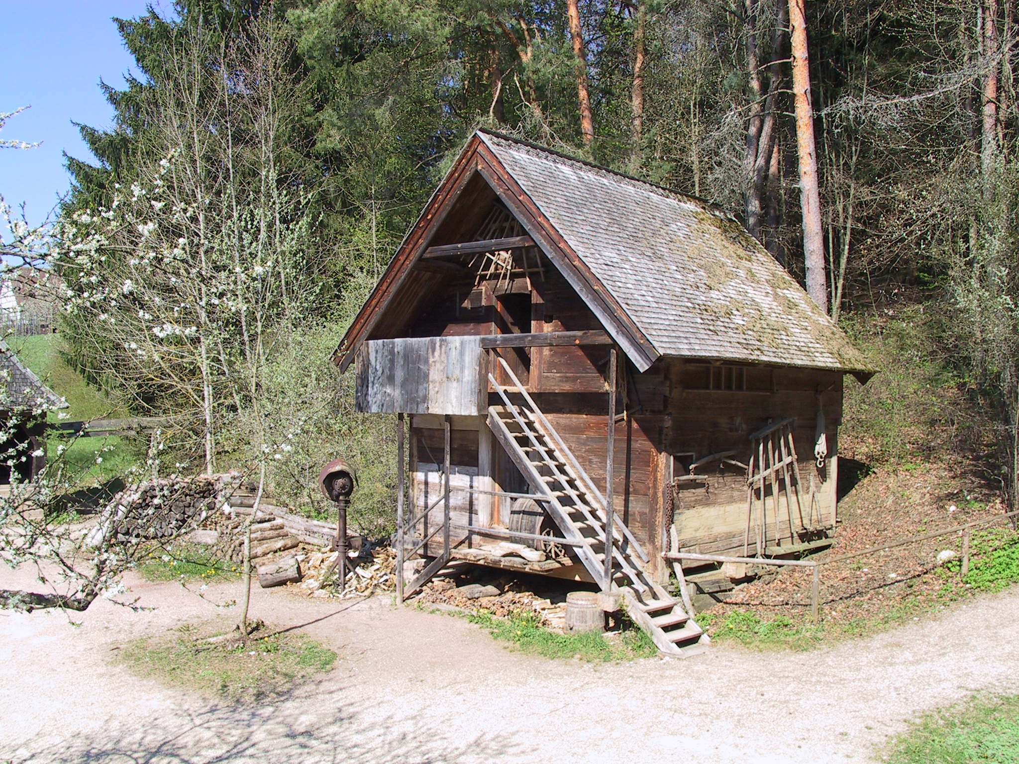 Foto: Kleiner Getreidespeicher aus Holz mit Schindeldach umgeben von Bäumen. Eine hölzerne Treppe führt zum oberen Eingang. Vor dem Gebäude liegen Holzscheite, an der Wand hängen alte landwirtschaftliche Geräte.