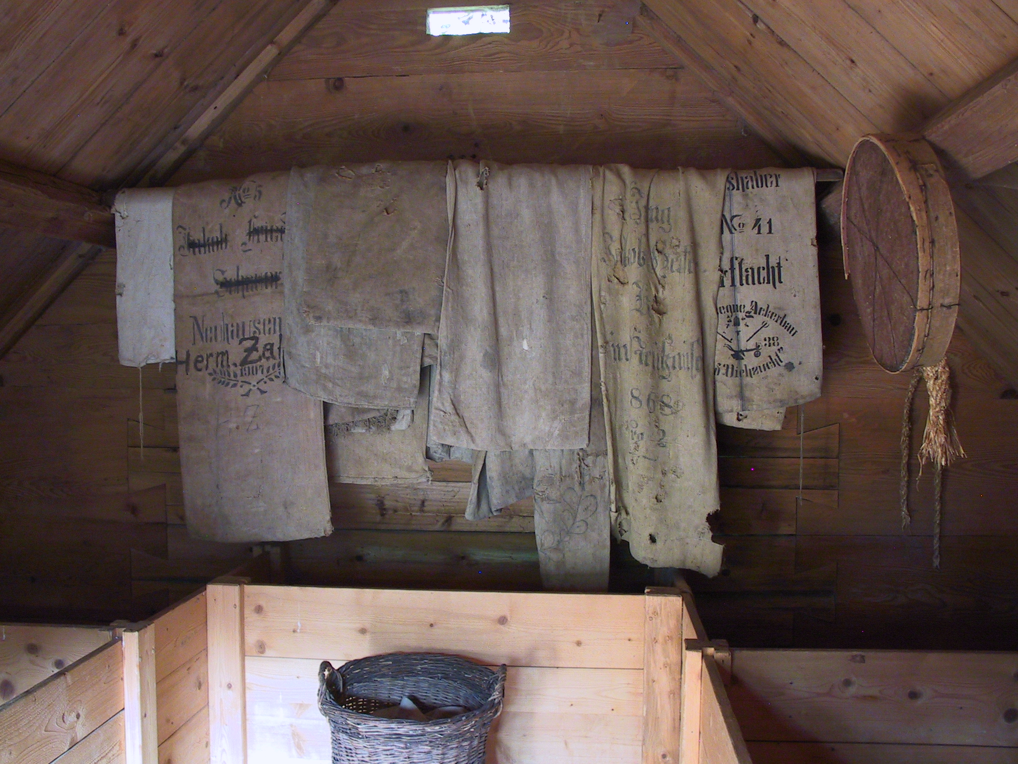 Foto: Innenansicht eines Getreidespeichers. An einem Balken hängen alte, bedruckte Getreidesäcke aus Leinenstoff. Rechts daneben ein Sieb und grobe Seile, unten ein geflochtener Korb.