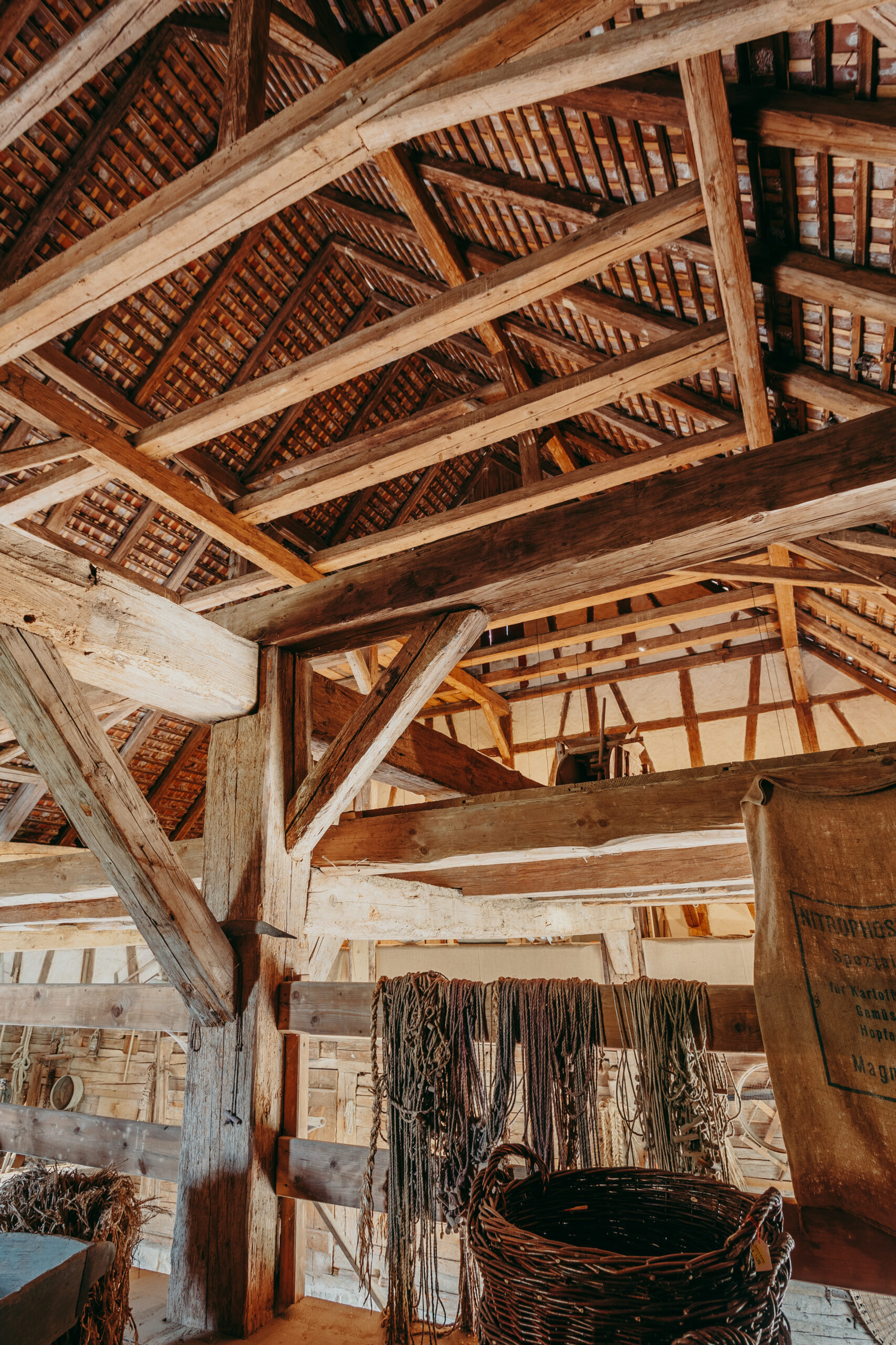 Foto: Blick auf den offenen Dachstuhl des Stalls mit Holzbalken und Dachziegeln. Im Vordergrund hängen Seile über einem Geländer, daneben ein geflochtener Korb und ein alter Jutesack.