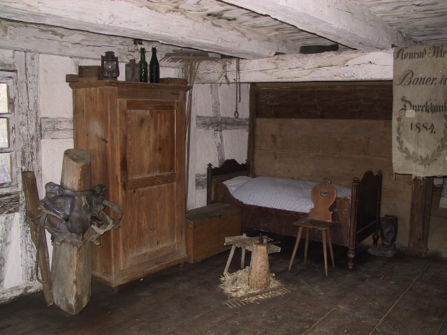 Foto: Historisches Bauernzimmer mit weiß gestrichenen Balken, einem Holzschrank, einer hölzernen Truhe, einem rustikalen Bett mit kariertem Bettzeug, einem Stuhl und einfachem Hocker auf Strohboden.