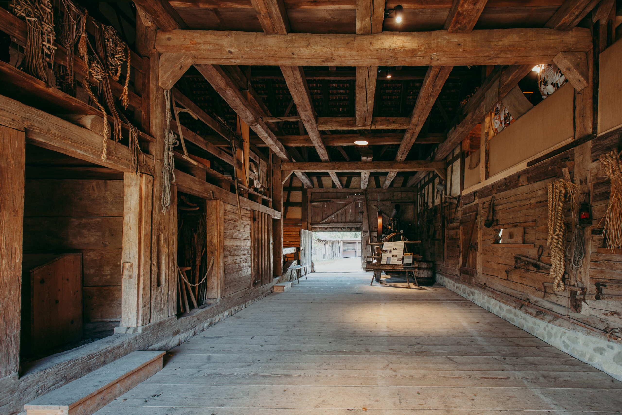 Foto: Innenraum des Stalls mit offenem Dachstuhl, Holzbalken, Seilen, Gerätschaften und Arbeitsmaterialien. Der Boden ist aus breiten Holzdielen. Am Ende des Stalls fällt Tageslicht durch das geöffnete Tor.