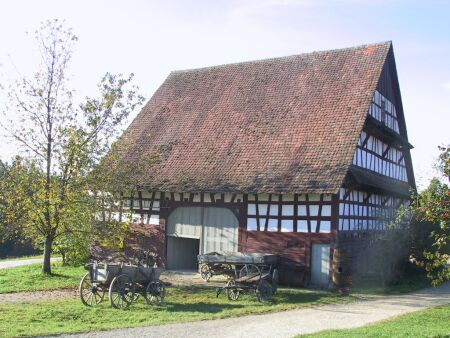 Foto: Außenansicht des Stalls in Fachwerkoptik mit steilem Ziegeldach. Davor stehen zwei alte Pferdewagen auf einer Wiese.