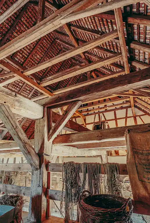 Foto: Blick auf den offenen Dachstuhl des Stalls mit Holzbalken und Dachziegeln. Im Vordergrund hängen Seile über einem Geländer, daneben ein geflochtener Korb und ein alter Jutesack.