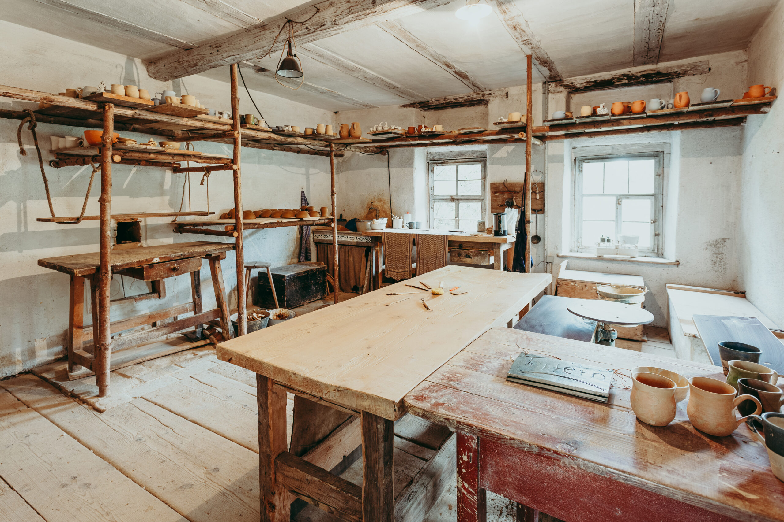 Foto: Innenansicht einer Töpferwerkstatt mit weiß gestrichenen Wänden, hölzernen Arbeitstischen und Regalen voller Keramikgefäße und Arbeitsmaterialien, zwei Fenster spenden Tageslicht.