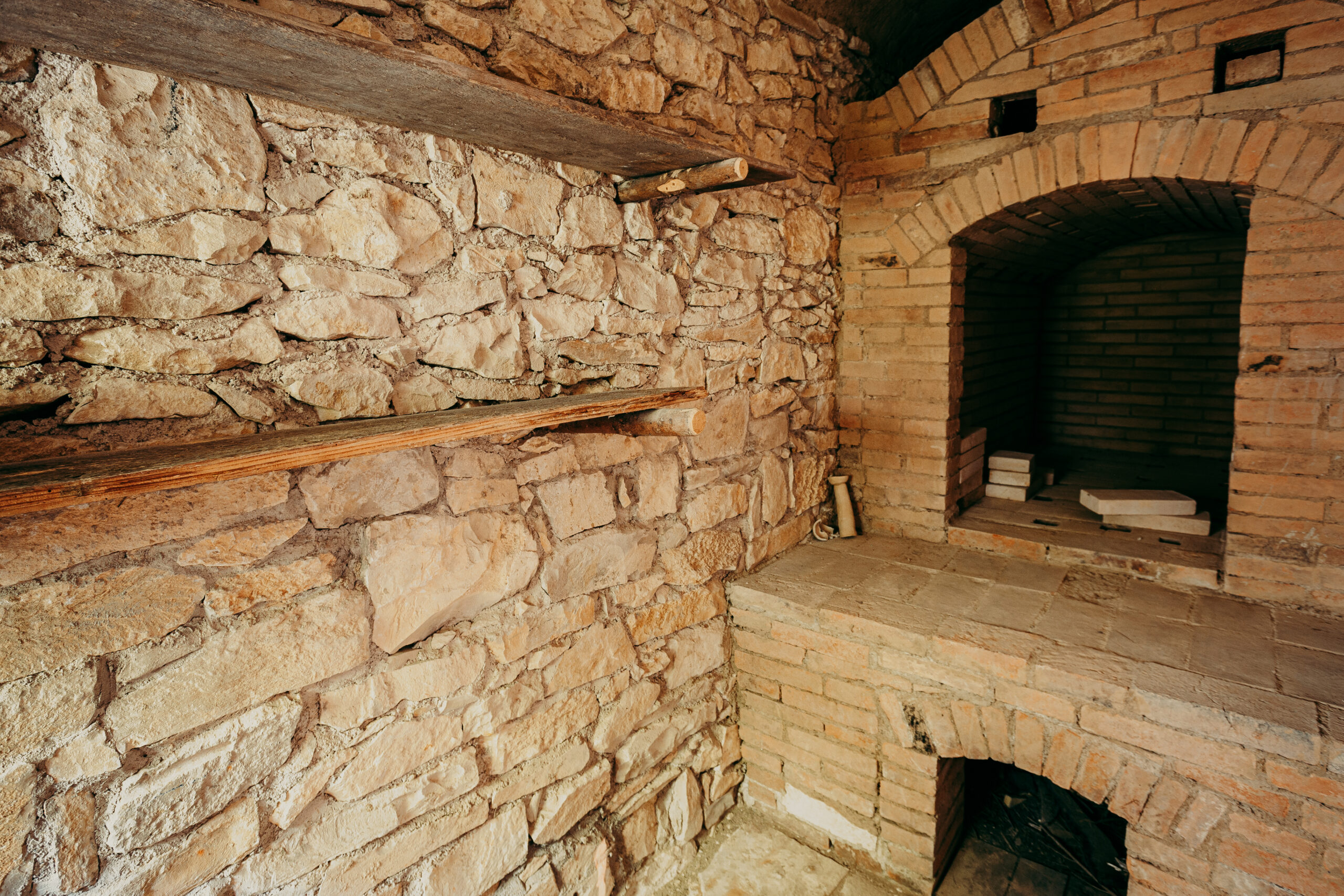 Foto: Innenraum mit unregelmäßig behauenen Natursteinwand an der zwei Holzablagen hängen und Backsteinofen mit gemauerter Feuerkammer.