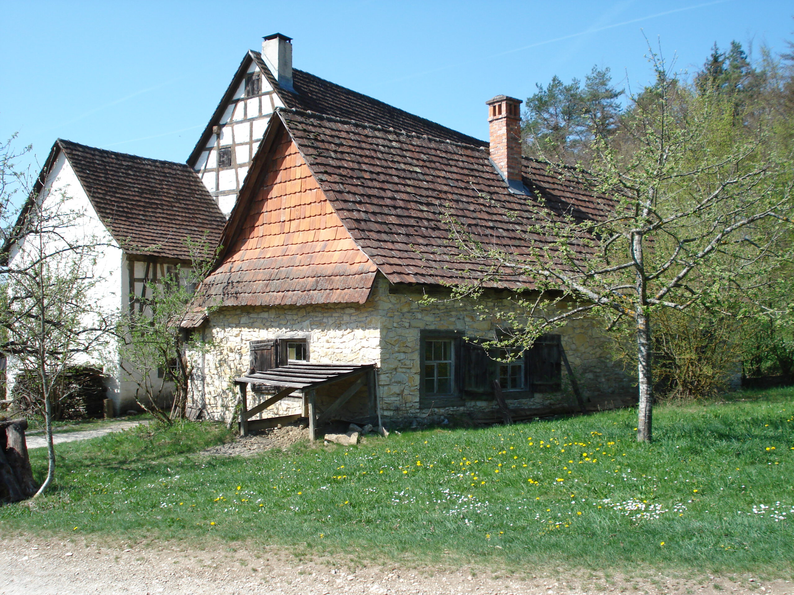 Foto: Außenansicht eines kleinen Hauses mit Natursteinmauerwerk und roten Dachziegeln, mit Holzläden an den Fenstern. Davor steht auf einer Wiese ein Baum.