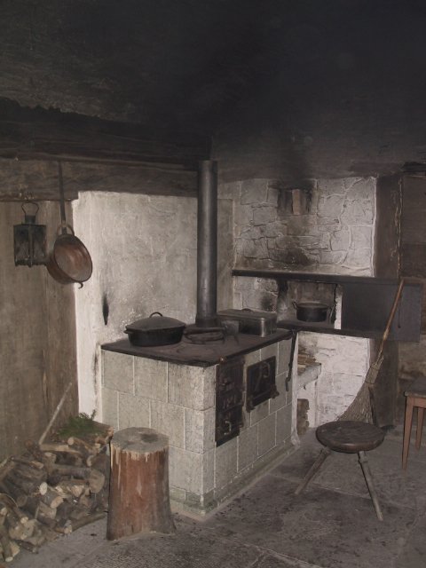 Foto: Historische Küche mit einem gemauerten Holzofen, auf dem mehrere schwarze Töpfe stehen. Holzvorräte und ein Hocker stehen auf dem dunklen Steinboden.