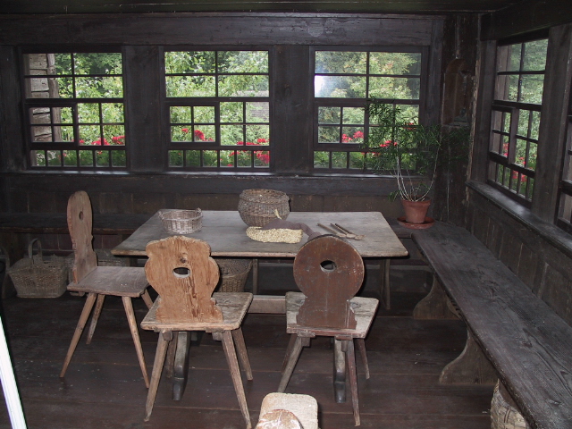 Foto: Dunkler, holzverkleideter Wohnraum mit einfachem Holztisch, drei Stühlen und einer langen Bank an der Wand. Auf dem Tisch liegen Körbe. Der Blick durch die Fenster offenbart die grüne Umgebung des Hauses und rote Blumen vor den Fenstern.