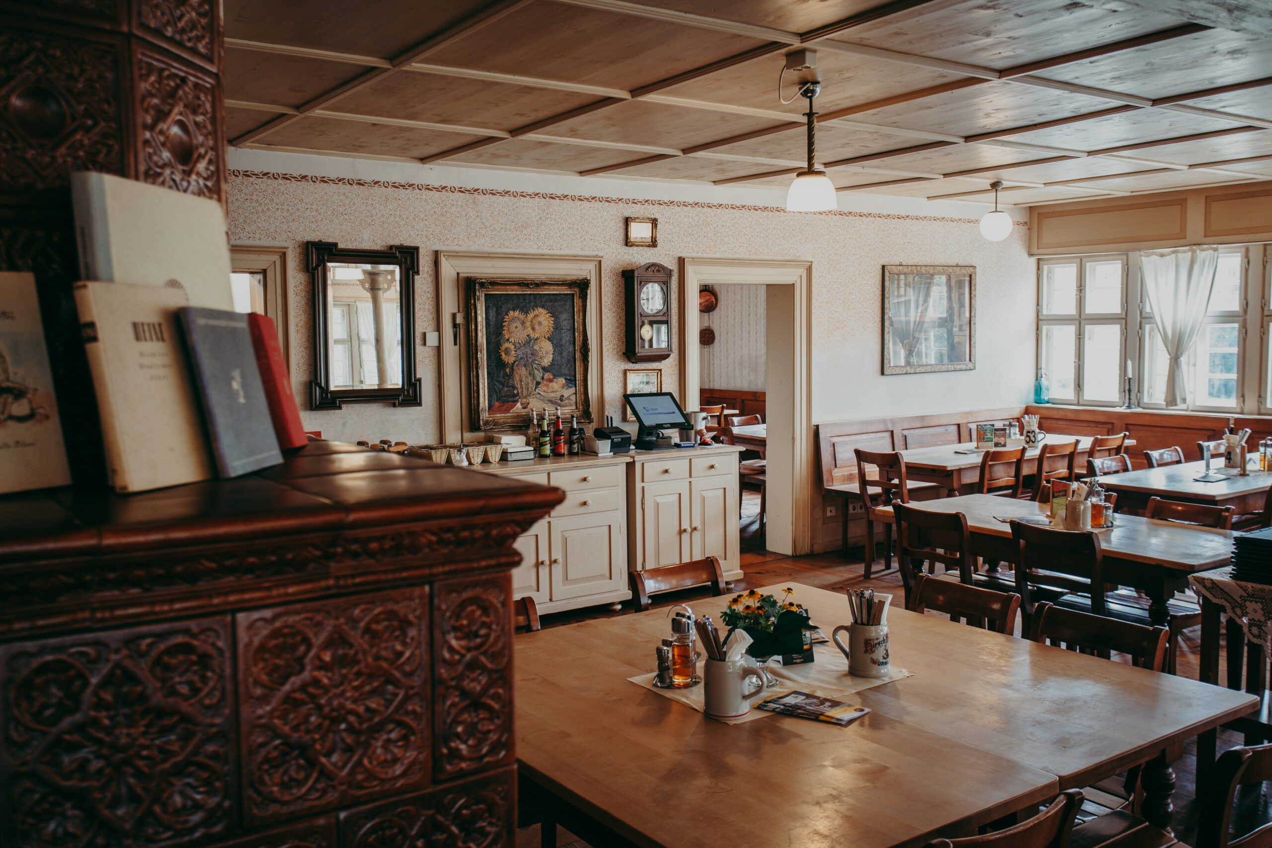 Foto: Innenraum eines traditionell eingerichteten Gasthauses mit Holzmöbeln, Kachelherd, dekorierten Wänden und eingedeckten Tischen, rechts große Fenster mit weißen Gardinen.