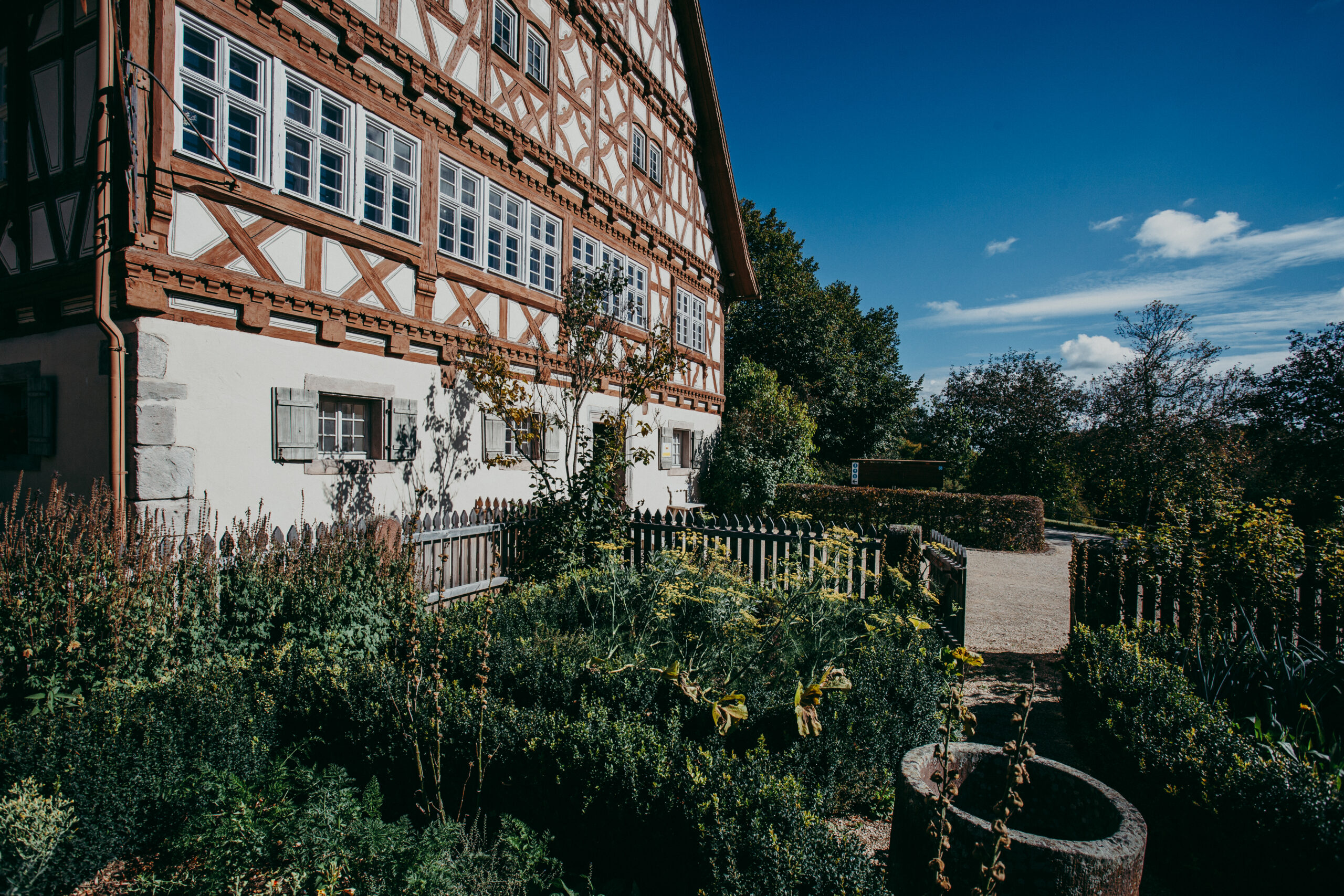 Foto: Ein kleiner, von einem Holzzaun abgegrenzter Garten vor einem großen Fachwerkhaus mit rot-braunem Gebälk.
