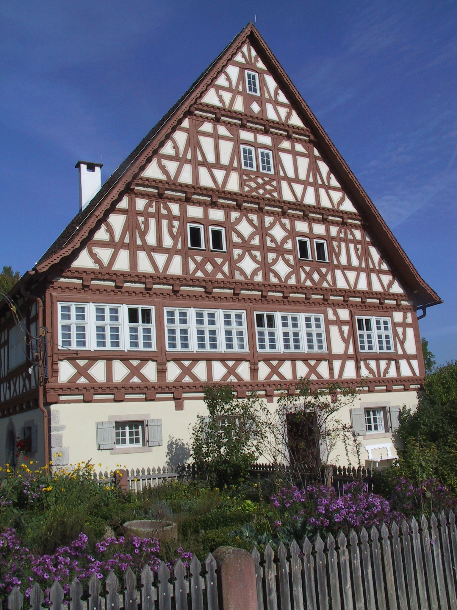Foto: Frontalansicht eines großen Fachwerkhauses mit rot-braunem Gebälk und weißen Fenstern, davor ein Garten mit bunten Blumen.