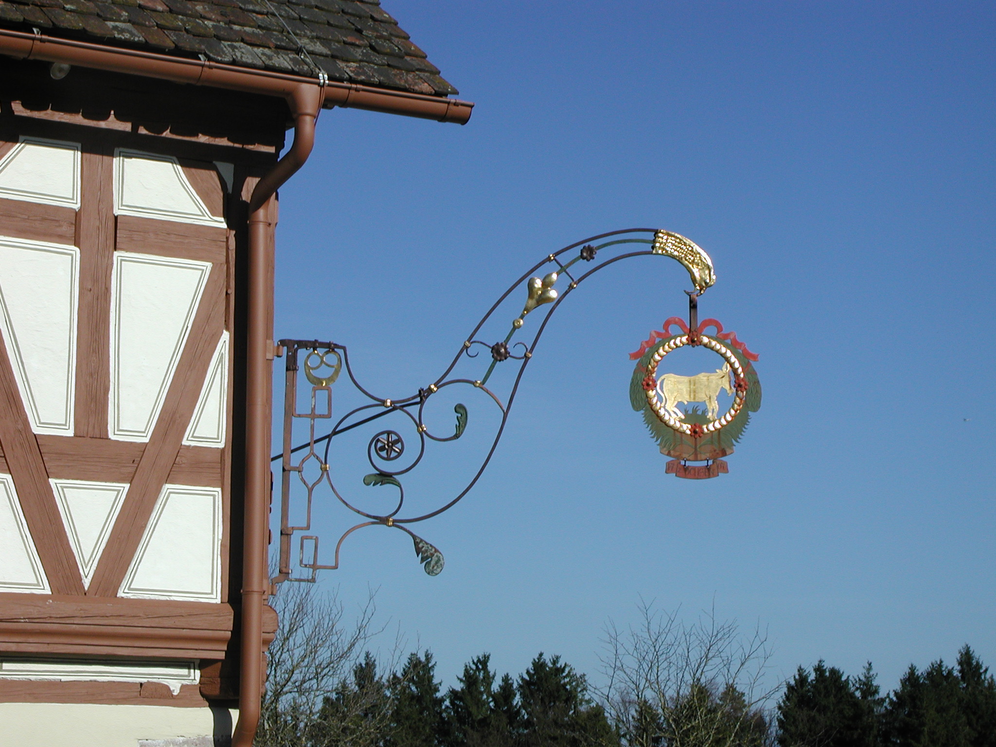 Foto: Schmiedeeisernes Wirtshausschild mit goldenen Verzierungen und einem Ochsen in der Mitte, befestigt an der Ecke des Gasthauses vor blauem Himmel.