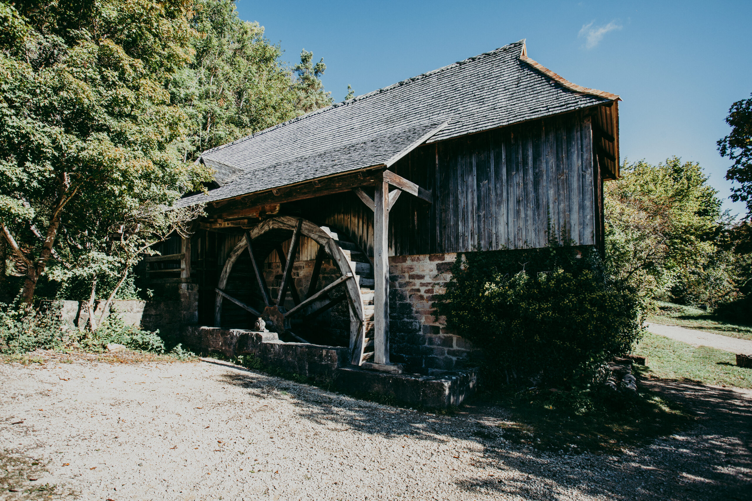 Foto: Sägewerk mit Wasserrad. Das Gebäude besteht aus Bruchsteinmauerwerk und Holzverschalung, das große hölzerne Wasserrad an der Wand des Sägewerks ist überdacht.