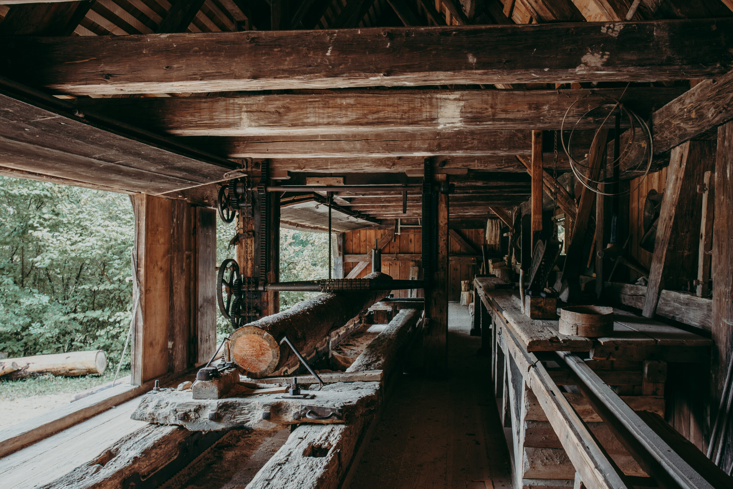 Foto: Innenansicht des Sägewerks. Ein Baumstamm ist auf einem Tisch eingespannt. Das Gebäude besteht vollständig aus Holz. Werkzeuge und Zahnräder sind sichtbar. Tageslicht fällt durch die offenen Seiten.