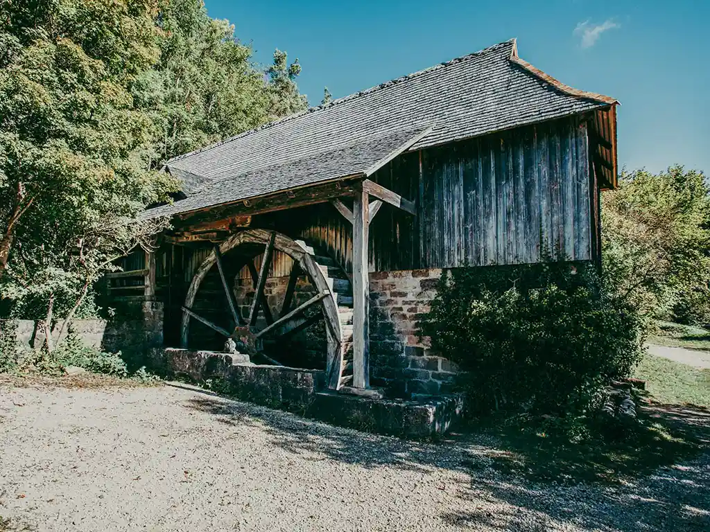 Foto: Sägewerk mit Wasserrad. Das Gebäude besteht aus Bruchsteinmauerwerk und Holzverschalung, das große hölzerne Wasserrad an der Wand des Sägewerks ist überdacht.