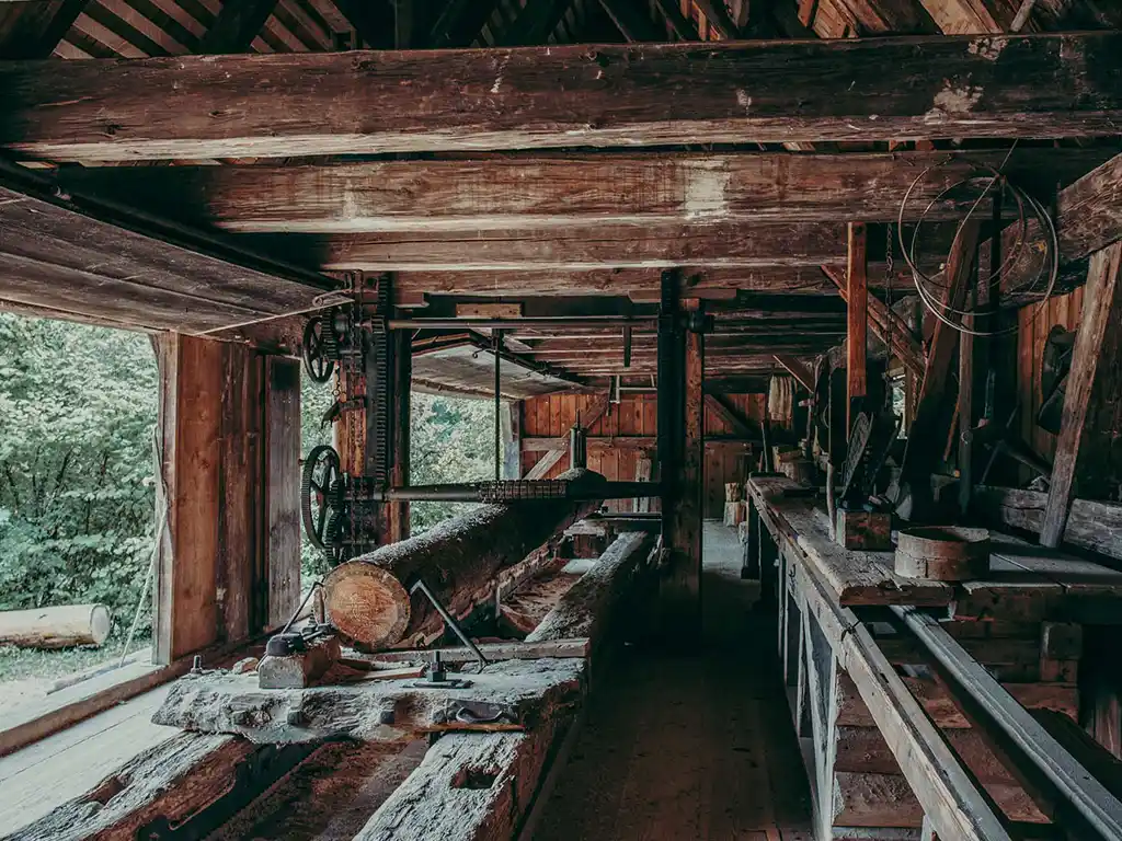 Foto: Innenansicht des Sägewerks. Ein Baumstamm ist auf einem Tisch eingespannt. Das Gebäude besteht vollständig aus Holz. Werkzeuge und Zahnräder sind sichtbar. Tageslicht fällt durch die offenen Seiten.