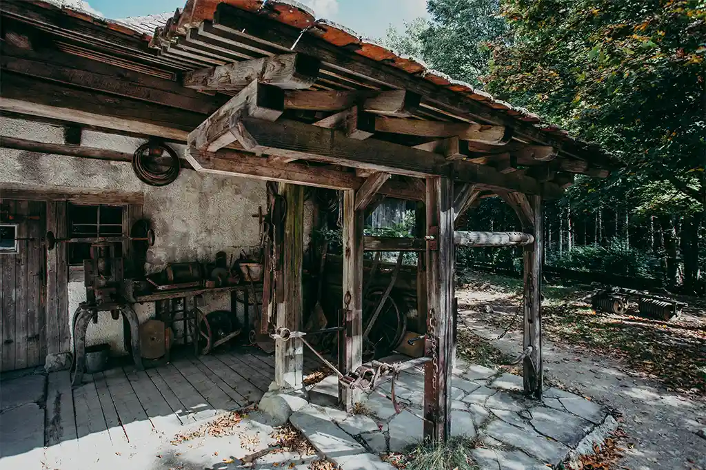 Foto: Historische Schmiede mit überdachtem Arbeitsbereich aus Holz. Unter dem Vordach stehen alte Werkzeuge und Geräte. Der Boden ist mit Holz und Steinplatten ausgelegt, umgeben von Wald und Laub.
