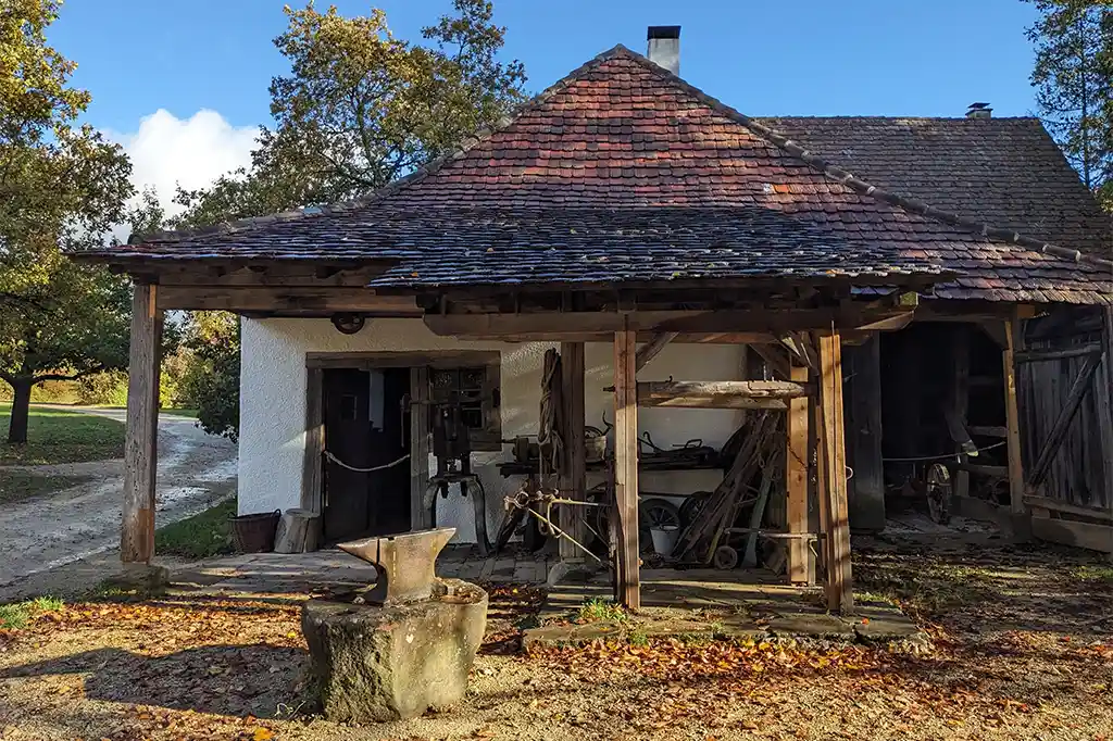 Foto: Gesamtansicht der historischen Schmiede mit rotem Ziegeldach. Vor dem Gebäude steht ein großer Amboss auf einem Steinsockel. Unter dem überdachten Arbeitsbereich sind verschiedene Werkzeuge und Geräte gelagert, umgeben von Herbstlaub.