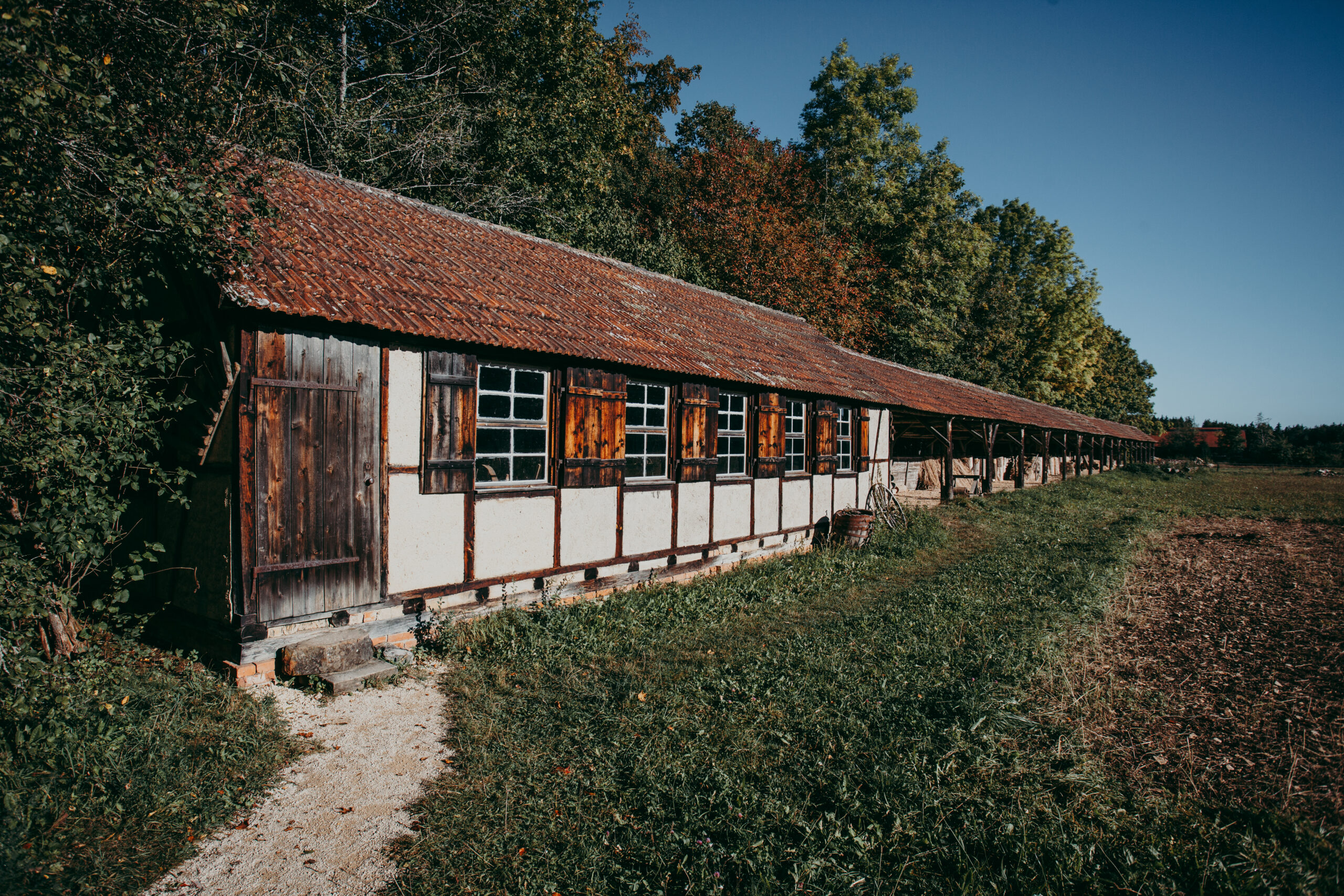 Foto: Langes Fachwerkgebäude mit weißen und braunen Wänden und roten Ziegeldachplatten. Die Fensterläden sind offen, daneben ist ein überdachter offener Bereich. Im Vordergrund ein Weg aus Gras und Kies, im Hintergrund Bäume.