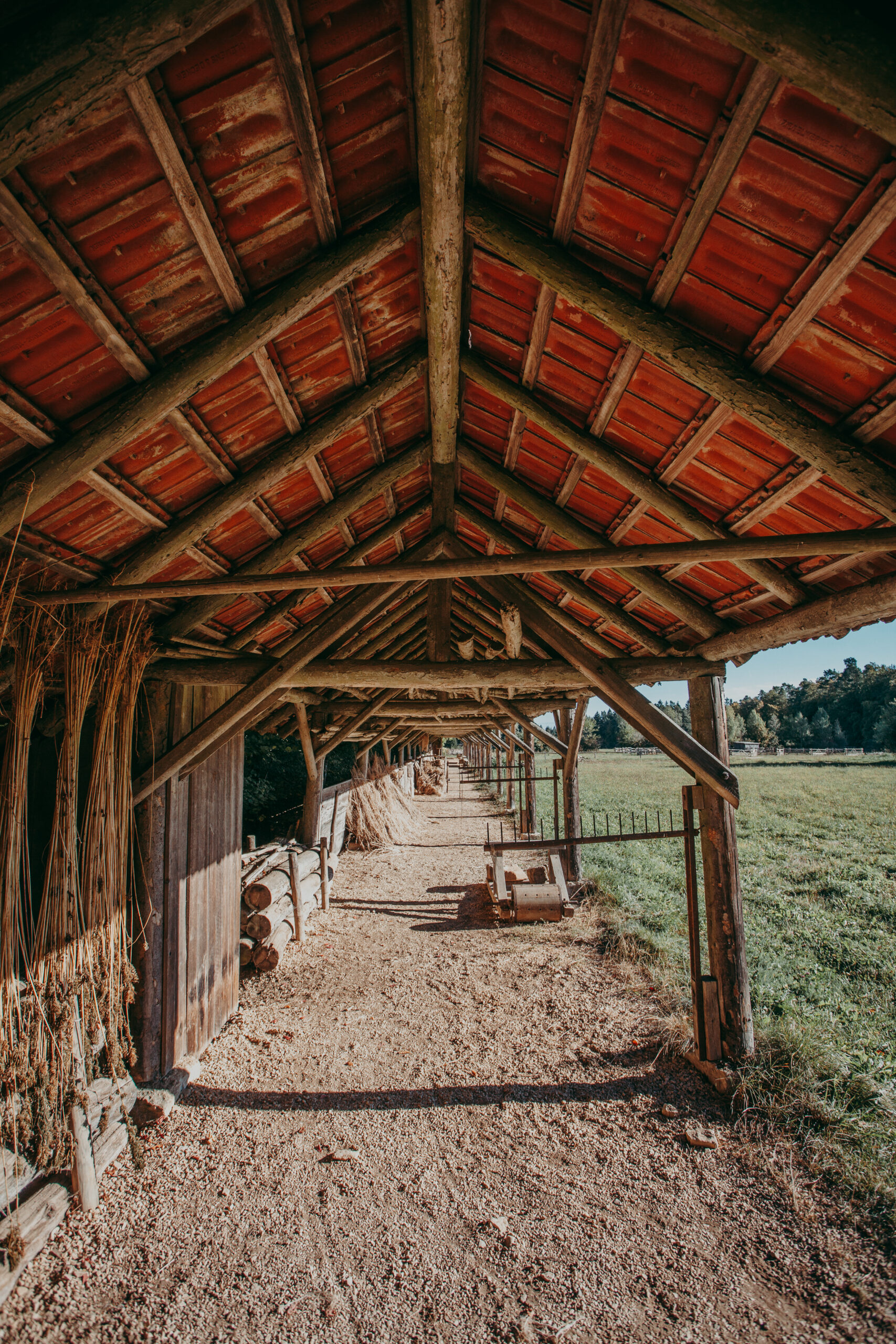 Foto: Blick in einen langen, offenen Gang unter einem Dach mit roten Ziegeln und Holzträgern. Links hängen Pflanzenbündel, rechts grenzt der überdachte Bereich an eine Wiese.
